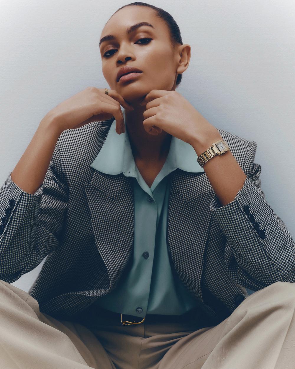 Elegant woman in houndstooth blazer, silk blouse, and trousers wearing wristwatch poses in indoor studio setting against white wall.
