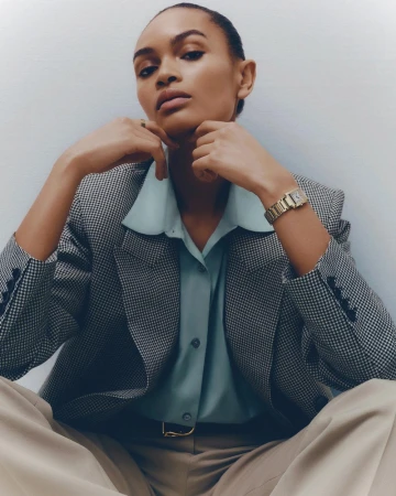Elegant woman in houndstooth blazer, silk blouse, and trousers wearing wristwatch poses in indoor studio setting against white wall.
