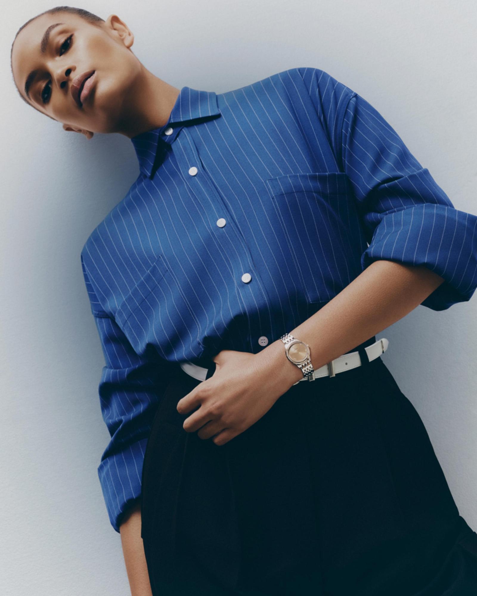 Elegant woman in a blue pinstripe shirt and black skirt poses in front of a white wall, showcasing a wristwatch and white belt.