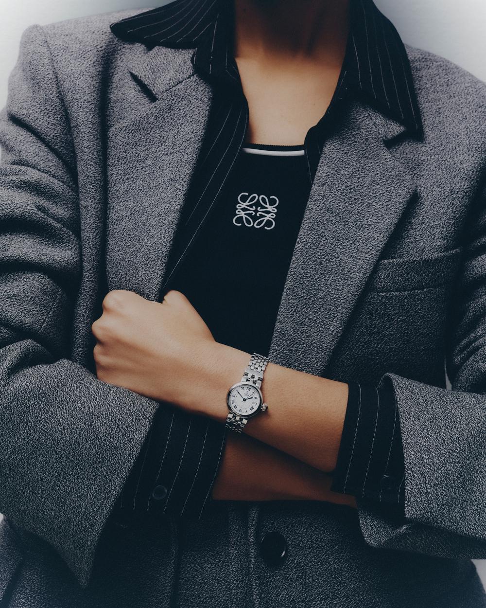 A woman in a gray blazer crosses her arms to display a silver watch on her left wrist, with a black shirt and gray pin-striped collared shirt.