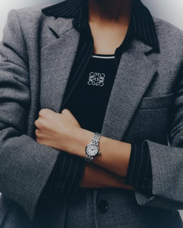 A woman in a gray blazer crosses her arms to display a silver watch on her left wrist, with a black shirt and gray pin-striped collared shirt.
