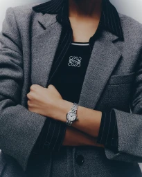 Close-up of a person wearing a grey tweed blazer, pinstripe shirt, and a silver wristwatch against a neutral white background