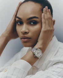 Black woman in a white pinstripe shirt wearing a silver luxury wristwatch with hands framing her face against a neutral background.