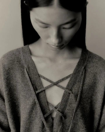 A young adult woman looks down, wearing a gray v-neck sweater with crisscross tie detail in a close-up, portrait-oriented fashion shot.