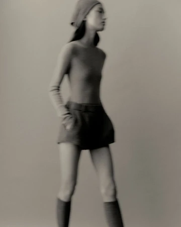 Monochrome portrait of a blurry woman in a beret, long-sleeve shirt, and shorts, walking in a neutral studio setting with soft, diffused light.