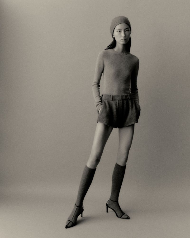 Full-length studio shot of a female model wearing a matching knit hat, long sleeve shirt, shorts, socks, and heels against a neutral backdrop.