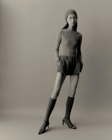 Full-length studio shot of a female model wearing a matching knit hat, long sleeve shirt, shorts, socks, and heels against a neutral backdrop.