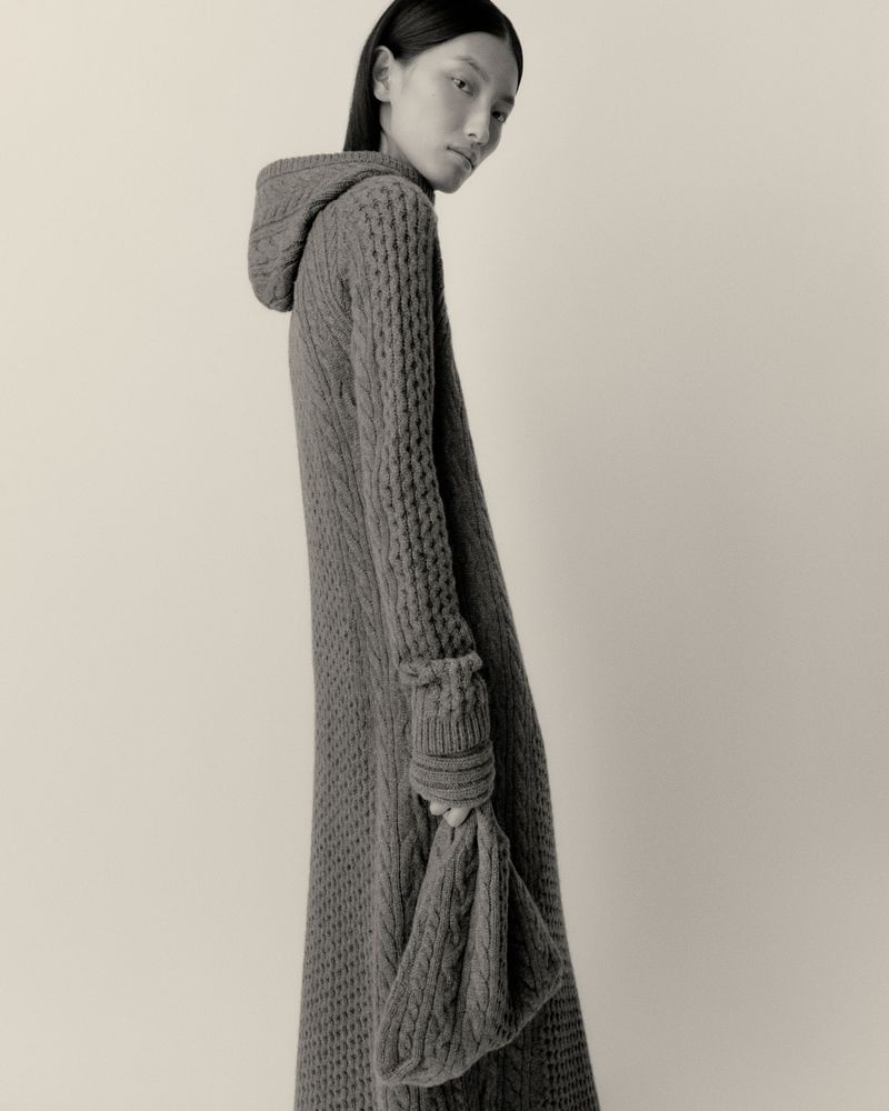 Monochromatic fashion portrait of an Asian adult female wearing a hooded, long-sleeved knit dress with a matching knitted bag.