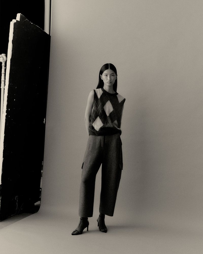 Indoor studio shot of a solitary young Asian woman in a patterned sweater vest, casual slacks, and heeled boots, posing elegantly.