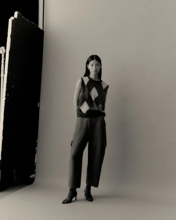 Indoor studio shot of a solitary young Asian woman in a patterned sweater vest, casual slacks, and heeled boots, posing elegantly.