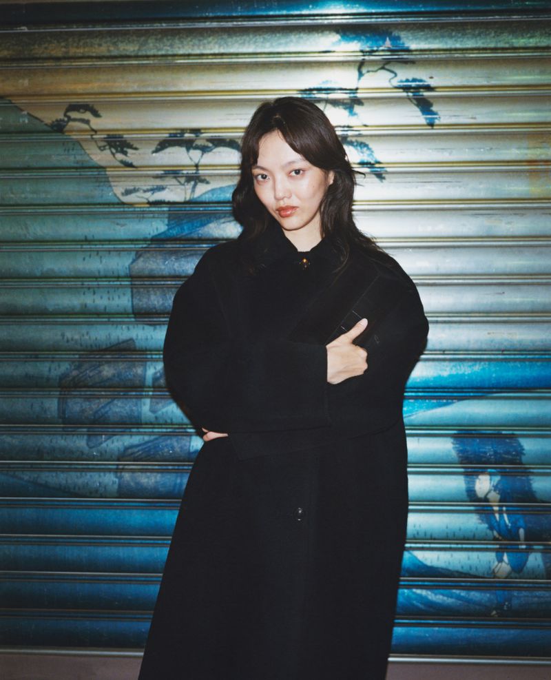 Young woman in a black coat stands confidently against a blue painted metal roll-up door backdrop in an urban setting, medium shot.