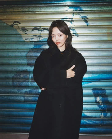 Young woman in a black coat stands confidently against a blue painted metal roll-up door backdrop in an urban setting, medium shot.