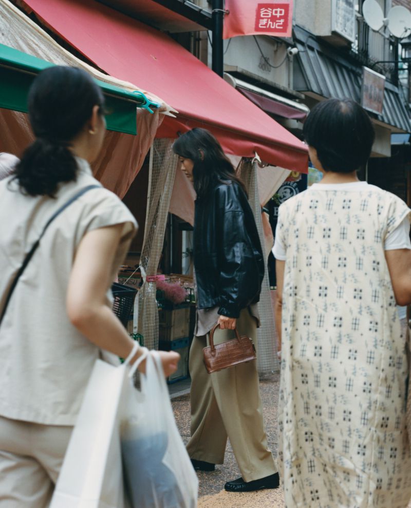 Three women walk on a city street lined with shops. One woman in the foreground carries a white plastic bag.