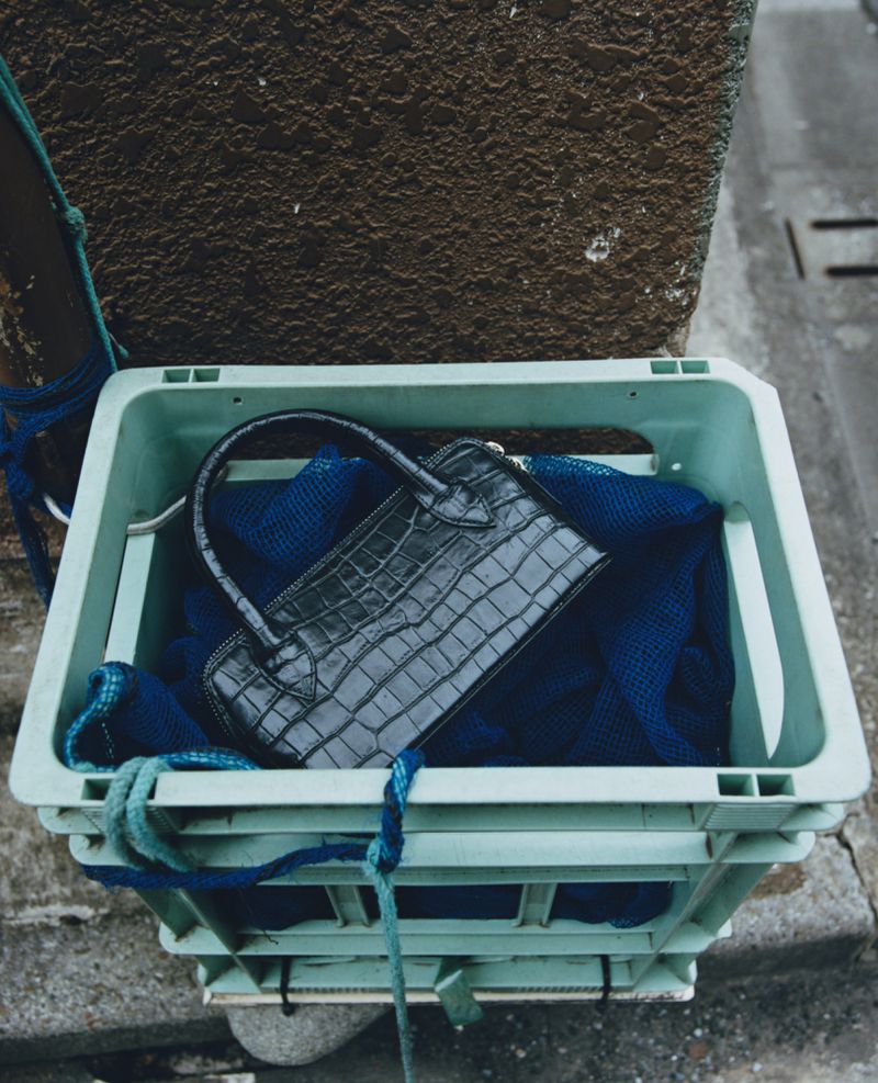 A black leather handbag with a crocodile pattern sits inside a green plastic crate outdoors with a dark blue cloth.