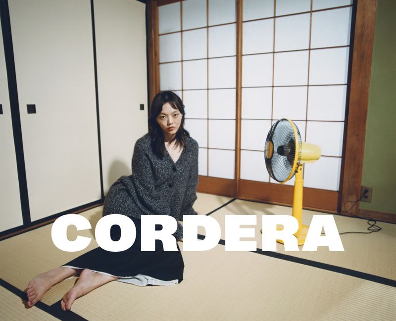 A young woman poses in an indoor setting with tatami mats, shoji screens, and a yellow fan, creating a minimalist and serene composition.