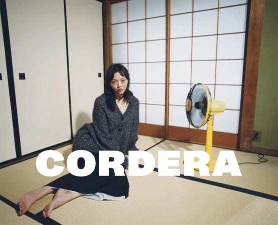 A young woman poses in an indoor setting with tatami mats, shoji screens, and a yellow fan, creating a minimalist and serene composition.