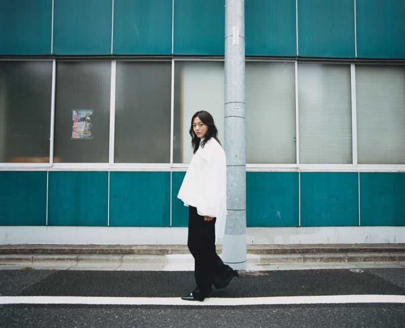 Woman in white shirt and black pants walks in front of teal building with large windows and a gray utility pole on a city street.