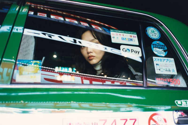Woman with dark hair looking out the window of a green taxi cab, stickers visible on the glass, indoor lighting, medium shot.