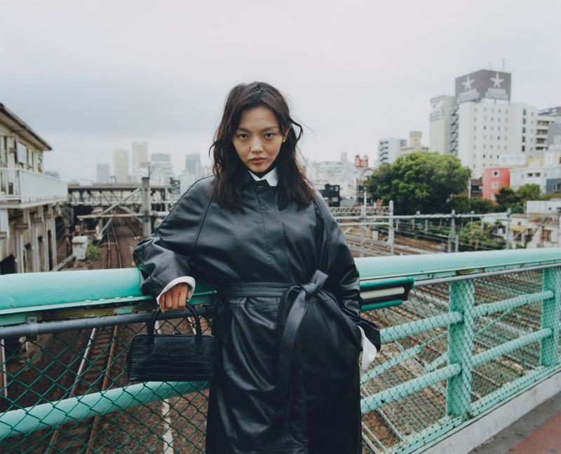 A woman in a black leather coat poses on an outdoor walkway with an alligator-skin purse; a Japanese city skyline is behind her.
