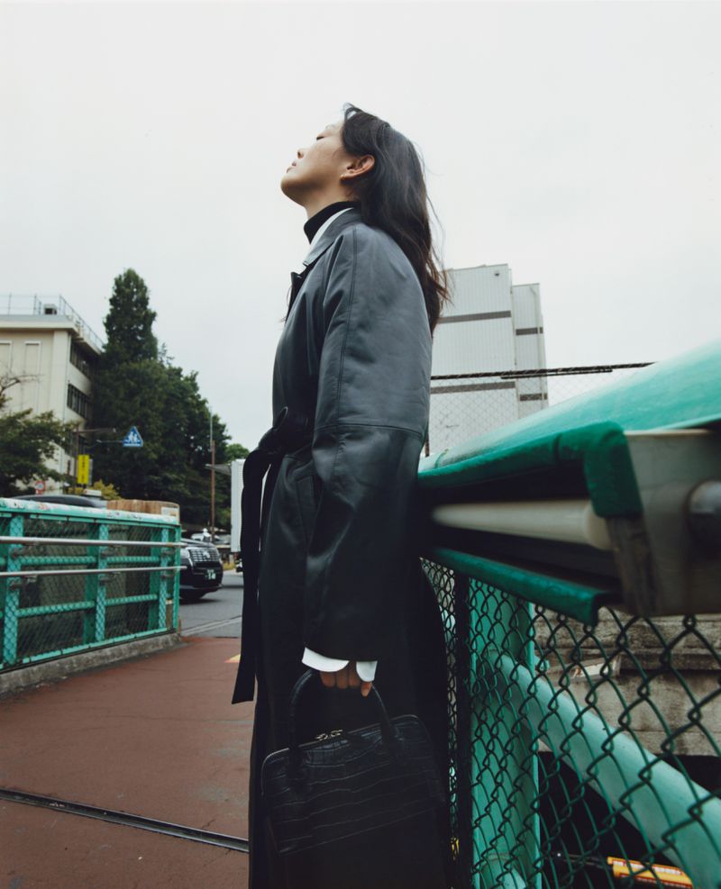 Side profile of an adult woman wearing a black trench coat and holding a black purse, standing outside in front of an urban cityscape.