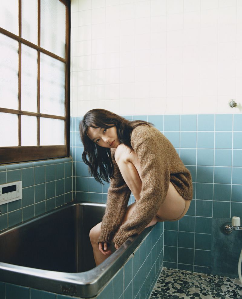 An Asian woman with long dark hair poses in a brown sweater while squatting on the edge of a metal bathtub in a tiled bathroom.