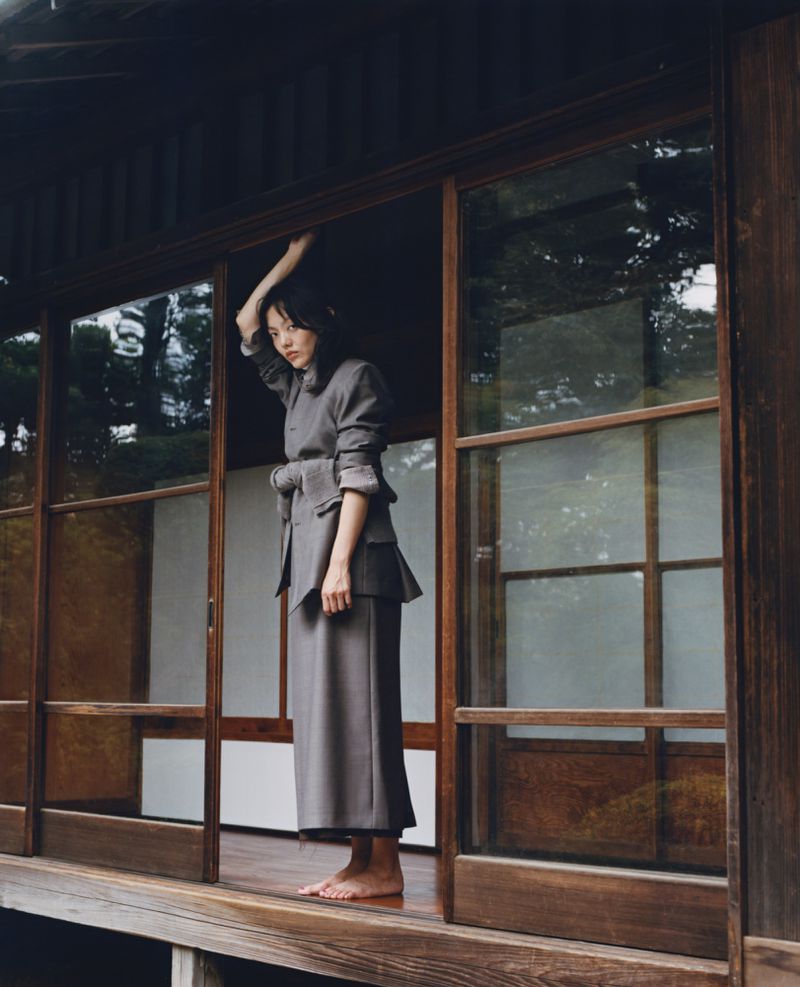 A woman in a gray suit stands barefoot in a traditional Japanese house with wooden frames and sliding glass doors, looking off-camera.