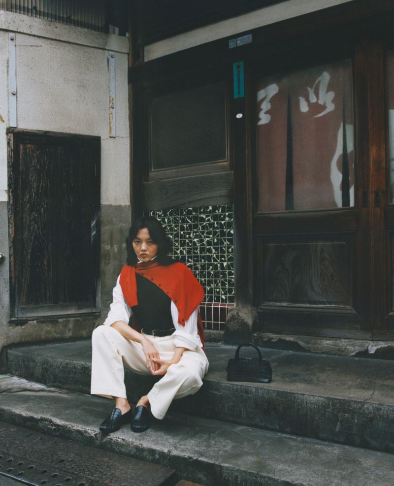 Stylish adult woman sitting on steps wearing a red shawl with a black bag in an urban setting next to a building with a dark entrance.