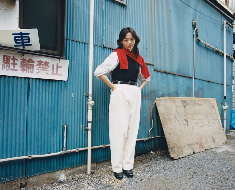 A stylish woman stands in front of a blue corrugated wall, wearing a black top, white trousers, and a red sweater around her shoulders.