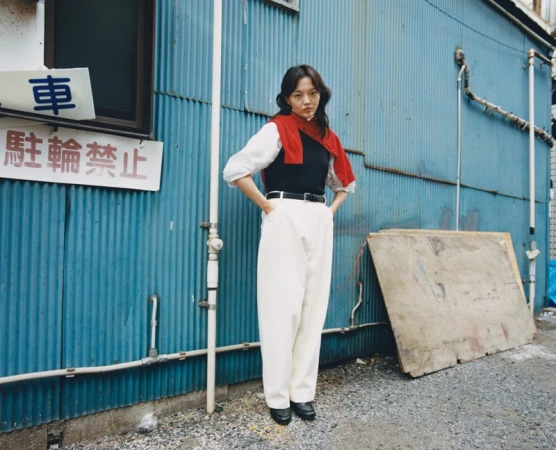 A stylish woman stands in front of a blue corrugated wall, wearing a black top, white trousers, and a red sweater around her shoulders.