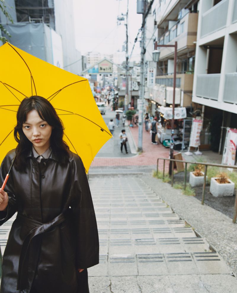 Woman in a black coat walking down stone steps with a bright yellow umbrella in an urban setting with buildings in the background.