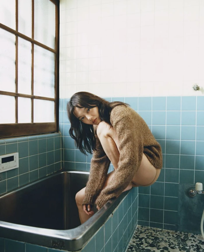 Rila Fukushima crouching on a bathtub edge wearing a brown knit sweater in a tiled bathroom for Cordera FW25