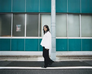 Rila Fukushima in a white blouse and black trousers standing on a street before a turquoise building for Cordera FW25