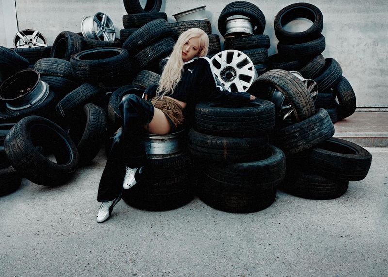 A woman in a black top and skirt sitting amongst a pile of black tires in a gray indoor area. Automotive fashion shoot.