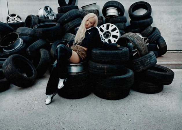 A woman in a black top and skirt sitting amongst a pile of black tires in a gray indoor area. Automotive fashion shoot.
