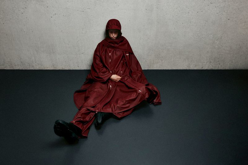 A person sitting on a dark floor wearing a maroon outfit with a cap and draped shawl against a grey textured wall in a studio setting.