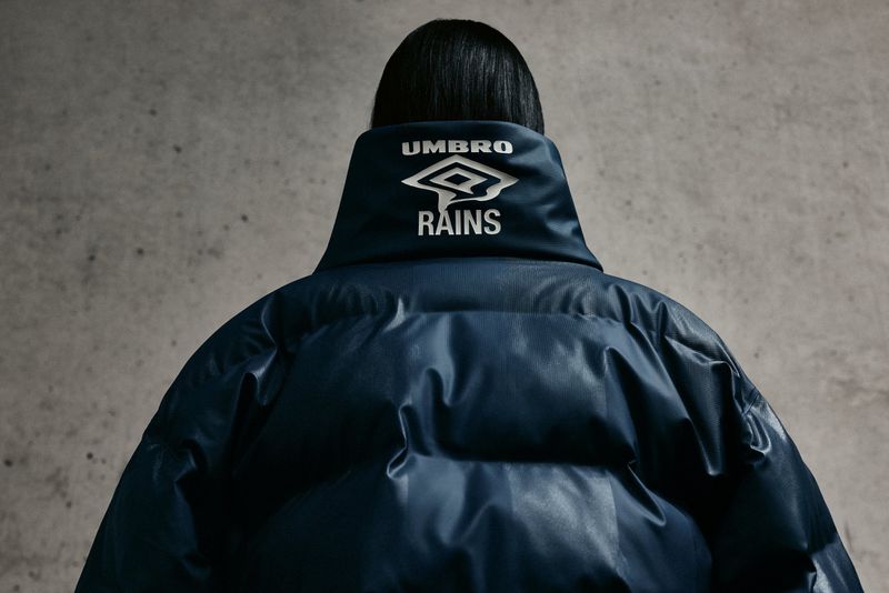 Back view of person in blue Umbro x Rains puffer coat against gray backdrop; fashion-forward outerwear and modern design.