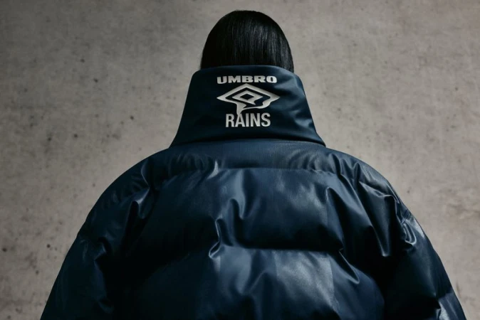Back view of person in blue Umbro x Rains puffer coat against gray backdrop; fashion-forward outerwear and modern design.