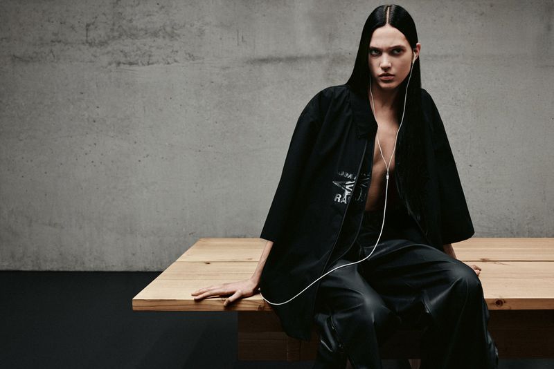 A person with long, dark hair sits on a wooden table in a black outfit with white headphones, against a grey wall for a studio fashion shoot.