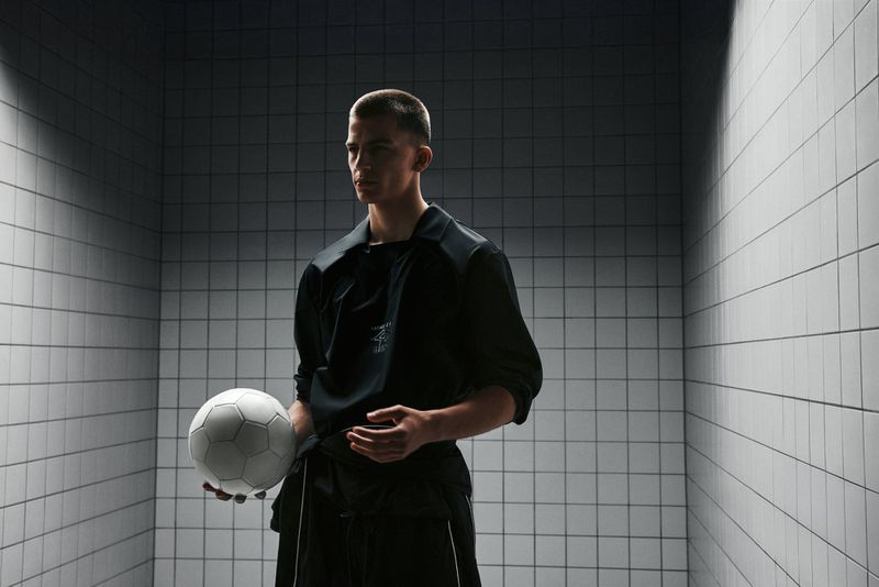Young adult male poses indoors holding a soccer ball, wearing a black jacket in a grey tiled room with overhead lighting.