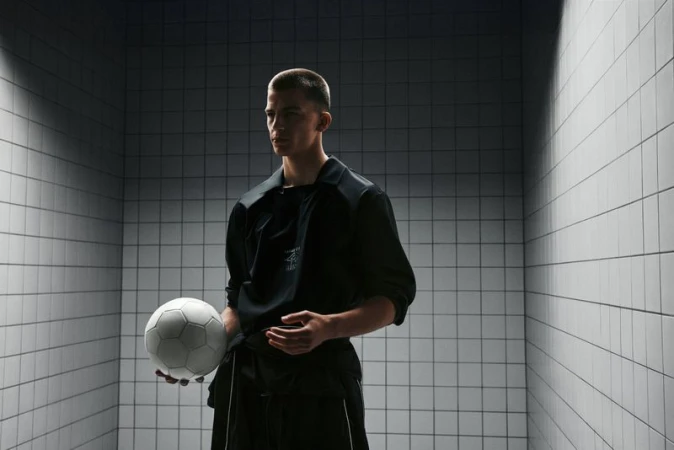 Young adult male poses indoors holding a soccer ball, wearing a black jacket in a grey tiled room with overhead lighting.