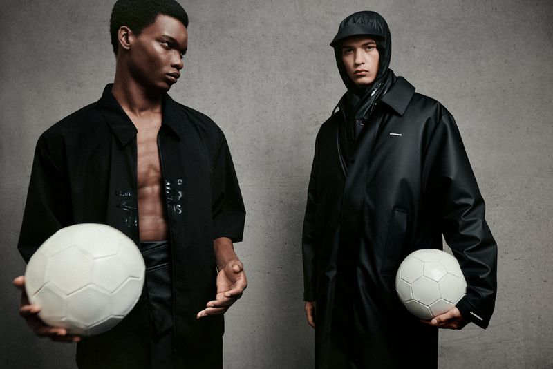 Two male fashion models pose in a studio setting with neutral background, each holding a white soccer ball. The models are wearing dark clothing.
