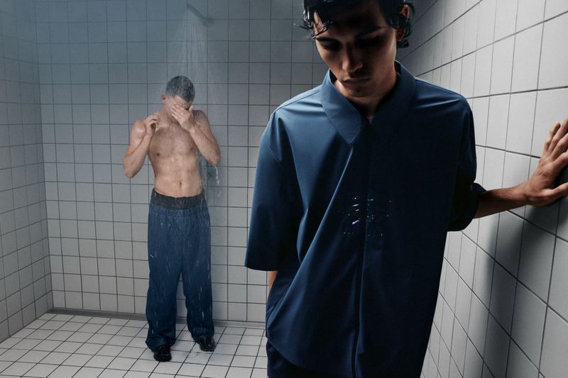 Two men in a tiled room: one under a shower, the other in a navy shirt leaning against the wall, creating a moody, fashion-forward scene.
