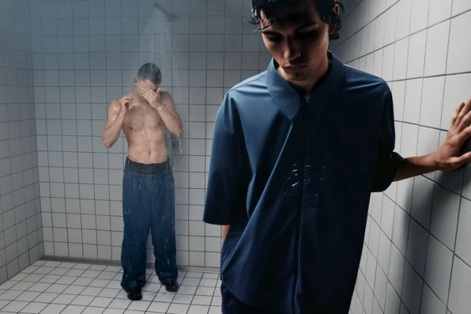 Two men in a tiled room: one under a shower, the other in a navy shirt leaning against the wall, creating a moody, fashion-forward scene.