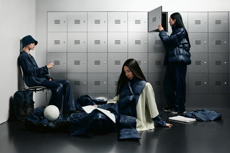 Three women in a locker room wear navy puffer jackets and shiny pants, one sits on the floor reading, one opens a locker.