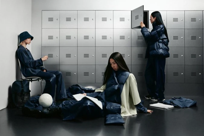 Three women in a locker room wear navy puffer jackets and shiny pants, one sits on the floor reading, one opens a locker.