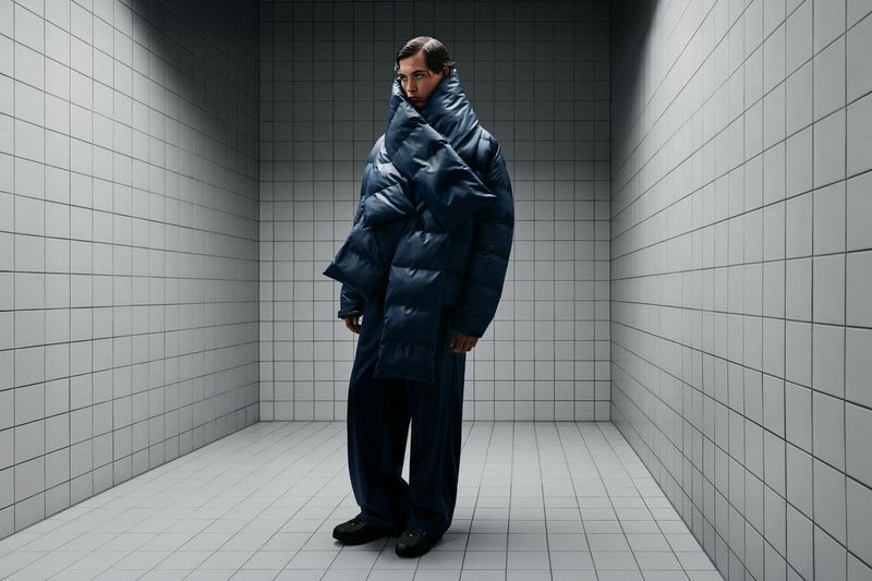 A slender adult male fashion model stands in a stark, tiled room wearing an oversized blue puffer jacket and pants, looking stoic.