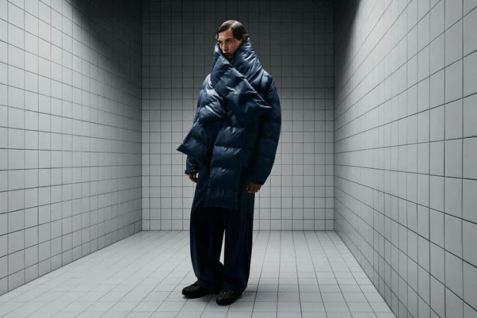 A slender adult male fashion model stands in a stark, tiled room wearing an oversized blue puffer jacket and pants, looking stoic.