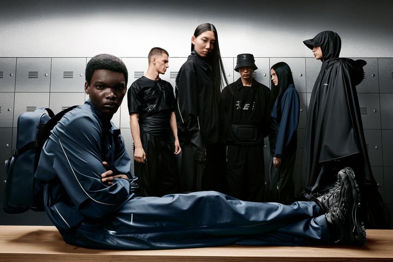 Group of fashion models in a studio pose against metal lockers, wearing coordinated athletic-inspired streetwear in dark tones.