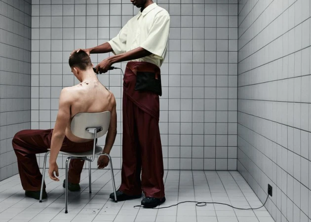 An adult male sits on a chair while another cuts his hair with electric clippers in a room with white tiled walls and floors.