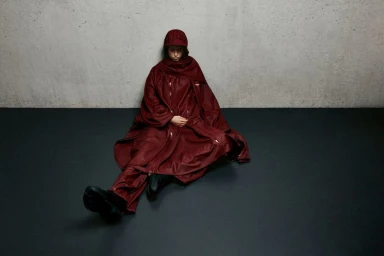 Model seated on a dark floor wearing a burgundy hooded waterproof coat and matching trousers against a concrete wall